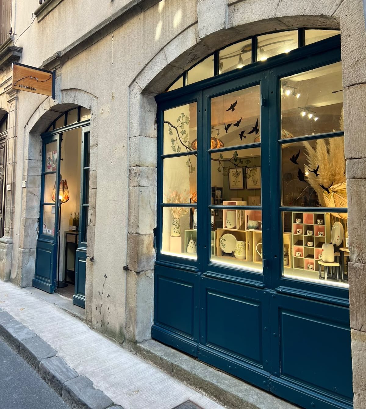 Devanture de M Céramique au 21 Grand Rue à Joyeuse : portes vitrées bleues sous une arche de pierre, oiseaux peints sur les vitres